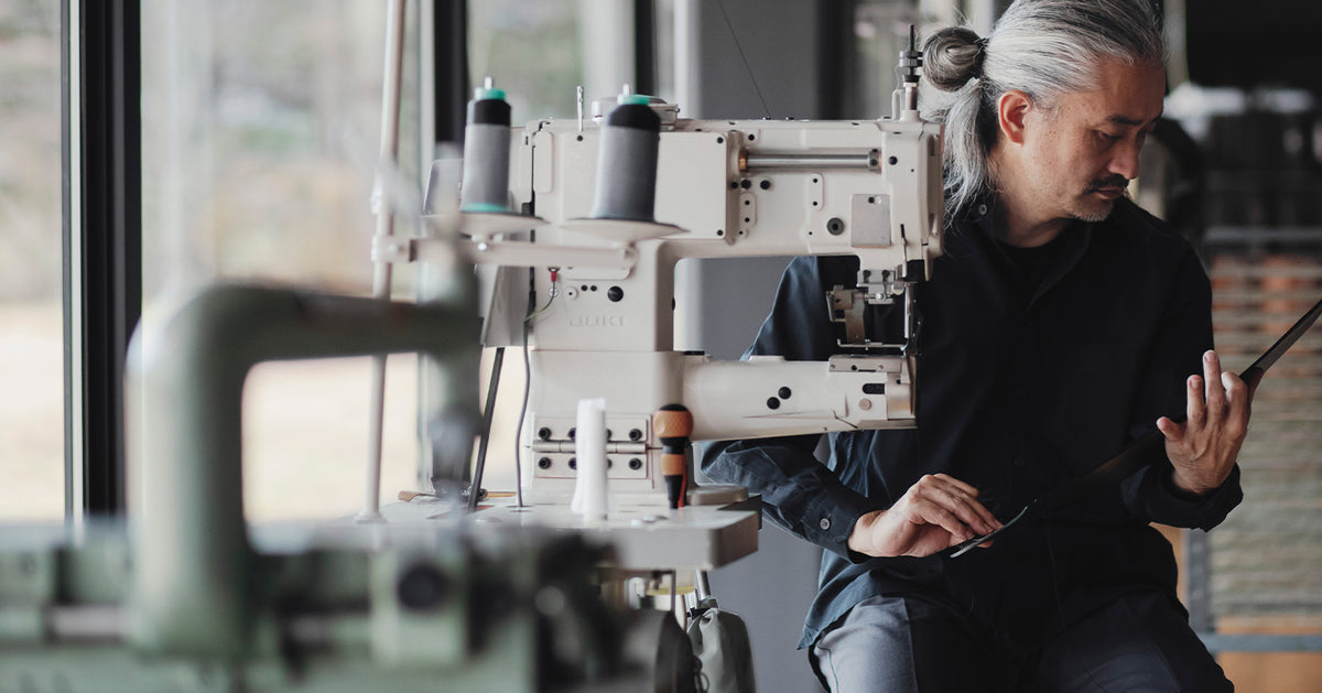 Isao Tamagawa of Craftsperson at Tsuchiya Kaban with long, grey hair tied back, wearing a dark shirt, sits beside an industrial sewing machine, holding a tablet and looking down at it.
