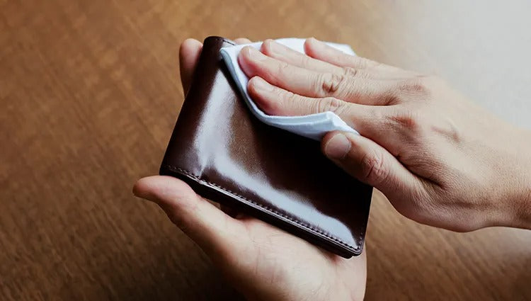 A close-up of two hands holding a shiny, dark brown leather wallet while a third hand wipes its surface with a white cloth.